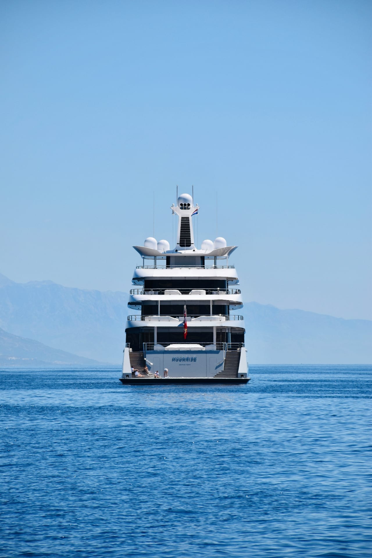 Multi-deck luxury superyacht anchored in calm blue waters with mountains in the background