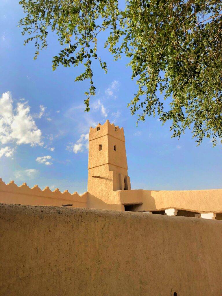 A historic tower in Diriyah, Saudi Arabia, framed by tree branches under a bright blue sky.