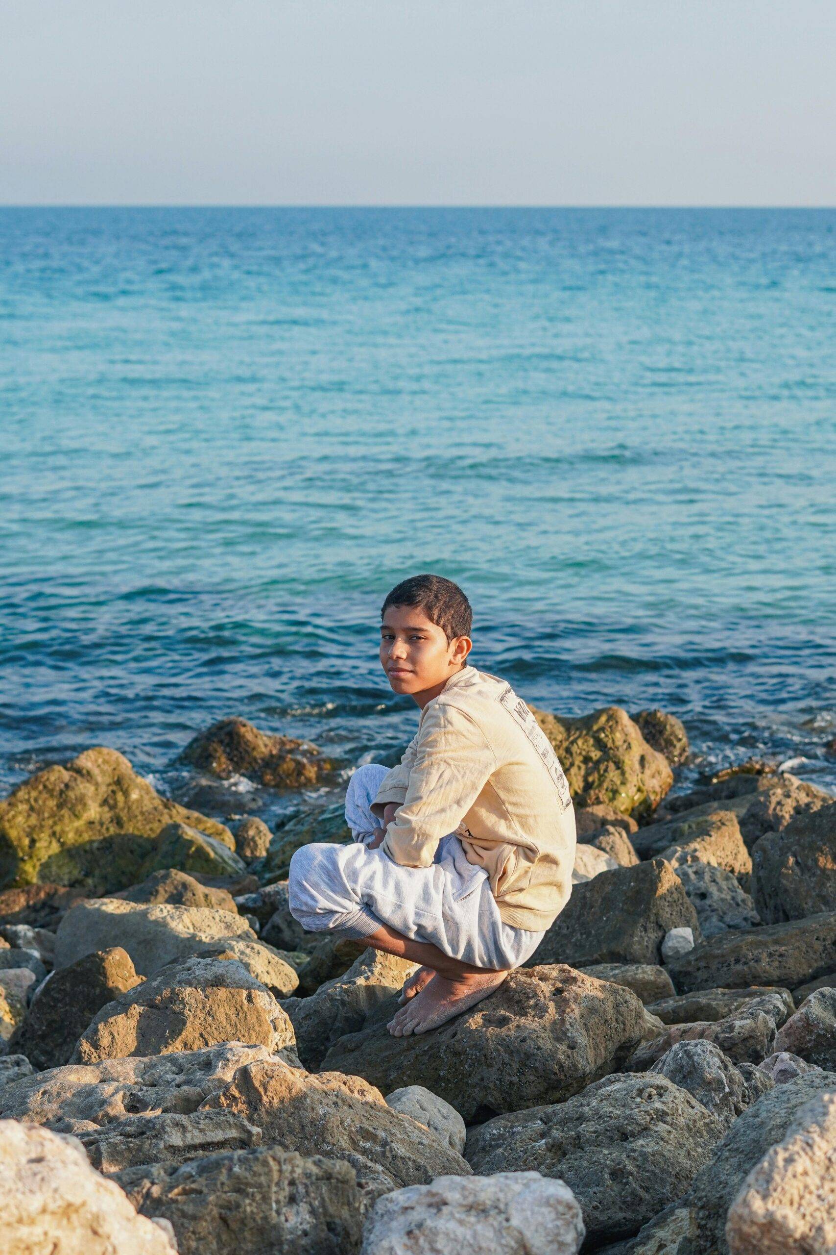 Teenager crouching on rocky beach by the sea in Khafji, Saudi Arabia during a sunny day.