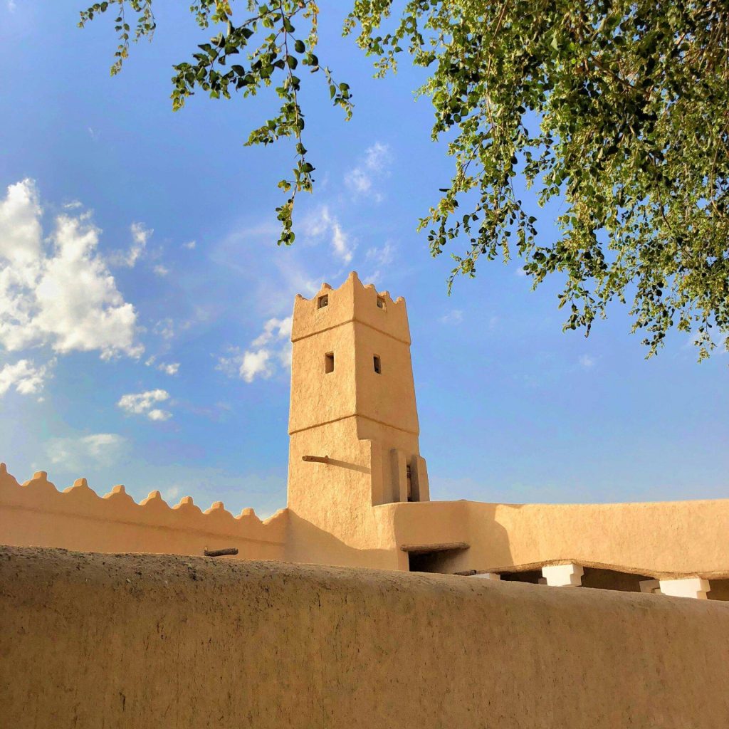 A historic tower in Diriyah, Saudi Arabia, framed by tree branches under a bright blue sky.
